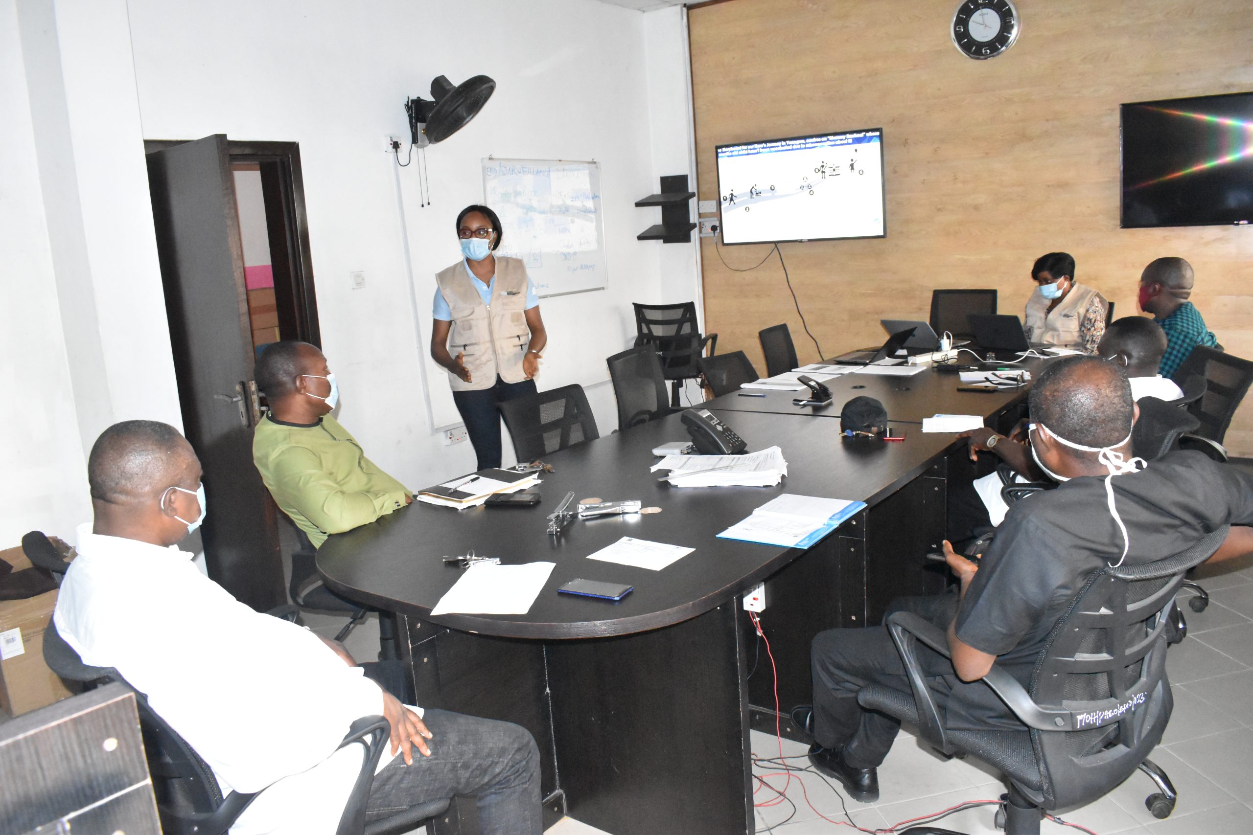 Group of 7 people who are part of the local government area (LGA) immunization team, all sitting around an oval desk with masks on in the middle of discussion with a presentation on display in the background